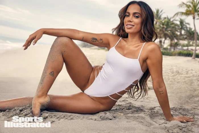 Anitta sits on the sand in a white one-piece with her knee bent and her arm propped on top of it.